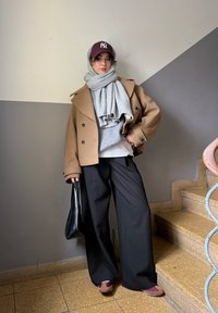 Brauner doppelt geclickter Mantel über grauem Pullover, schwarze weite Hose, grauer Schal und bordeauxfarbene Kappe, hält eine schwarze Tote-Bag, auf Treppen.