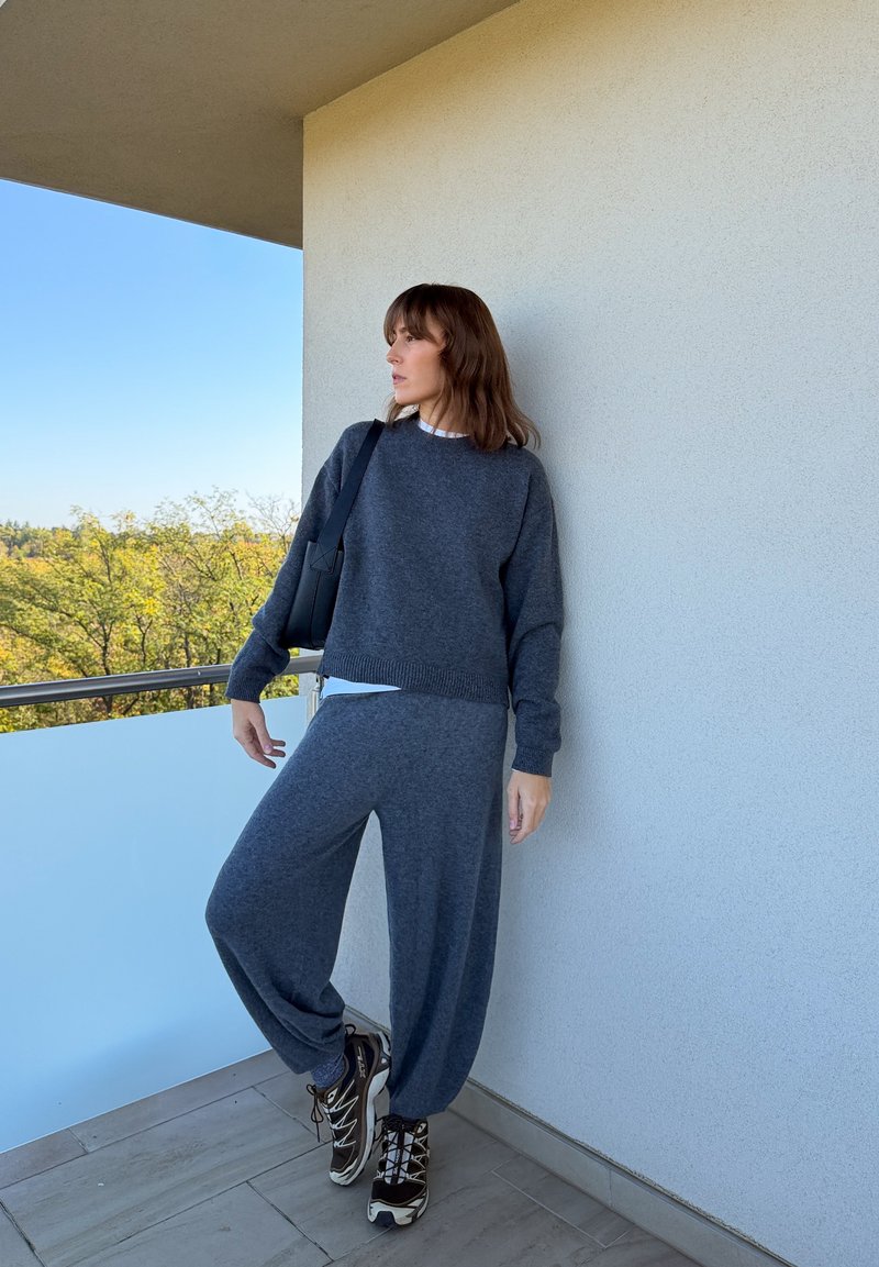 Une femme vêtue d’un pull en maille et d’un pantalon gris anthracite, de baskets et d’un sac à bandoulière se tient sur un balcon et regarde les arbres et le ciel bleu.