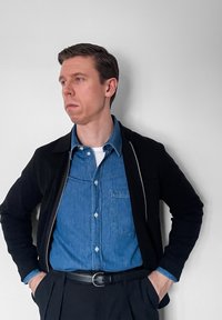 Homme en veste noire par-dessus une chemise en jean bleue et un pantalon noir, debout les mains dans les poches, regardant vers la gauche devant un mur uni.