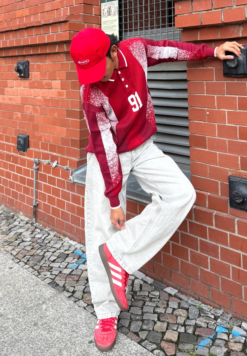 Rotes Polohemd mit langen Ärmeln, weißen Akzenten und der Nummer 91, kombiniert mit hellgrauer, weiter Jeans und roten Turnschuhen mit weißen Streifen. Ziegelsteinmauer im Hintergrund.