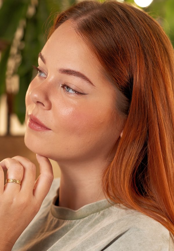 Profiel van een vrouw met lang, steil kastanjebruin haar, gekleed in een lichtgrijs T-shirt. Ze heeft een gouden ring om haar vinger en een zachte huid.