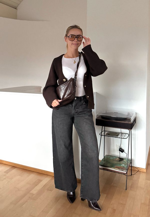 Mujer con gafas de sol grandes, cárdigan oscuro, camiseta blanca, vaqueros de pierna ancha y que sostiene un bolso cruzado de cuero oscuro en el interior.