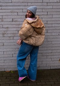Beige puffer jacket with a hood, pink sweater underneath, light blue wide-leg jeans, and pink shoes. Grey knitted beanie, small pink bag.