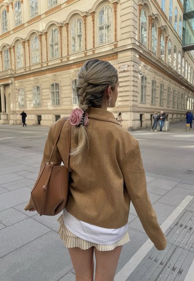 Chaqueta de cuero marrón con una textura suave, una falda plizada ligera y un bolso marrón. El cabello está peinado en una trenza con un accesorio floral rosa.