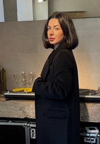 Fekete gyapjúkabát, középhosszú, egysoros gombolású kivitel. A háttérben egy lemezjátszó sárga tányérral, ezüst gyertyatartó és vinyl lemezek láthatók.