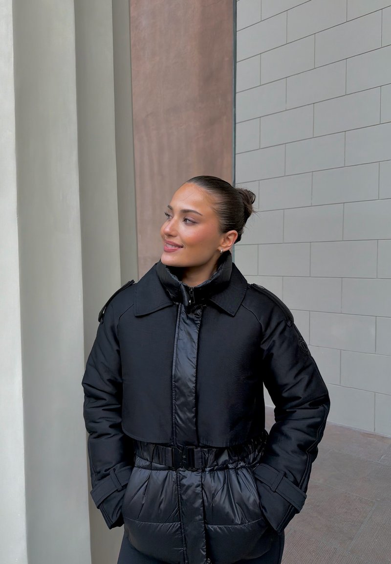 Giacca imbottita nera con design corto, colletto ampio e vita con cintura. Presenta una texture lucida e tasche anteriori.