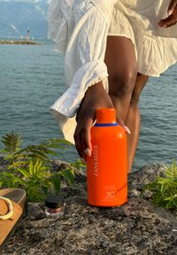 Orange solkrämflaska märkt "Lancaster Velvet Milk SPF 30" bredvid en liten svart burk, placerad på en stenig yta med grön vegetation.