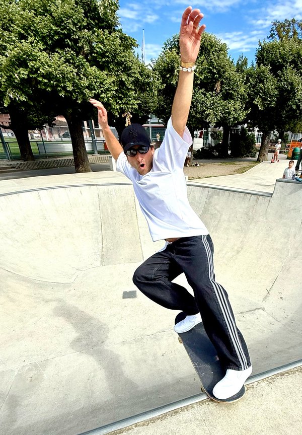 Un homme portant une casquette noire et des lunettes de soleil fait du skateboard en montant le mur courbe d'un bol en béton dans un skatepark par une journée ensoleillée.