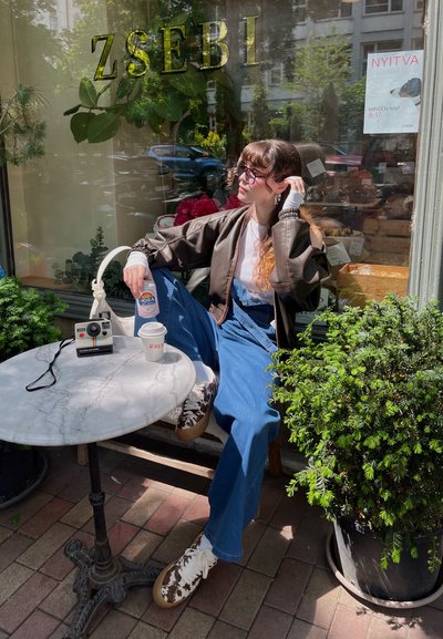 Chaqueta bomber marrón, camiseta blanca, overoles azules y zapatillas estampadas, sentado en una mesa de mármol con una cámara vintage y bebidas.