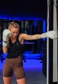 Boxeur portant des gants blancs et un haut noir sans manches, avec un short marron taille haute, lançant un coup de poing dans une salle de sport éclairée en bleu.