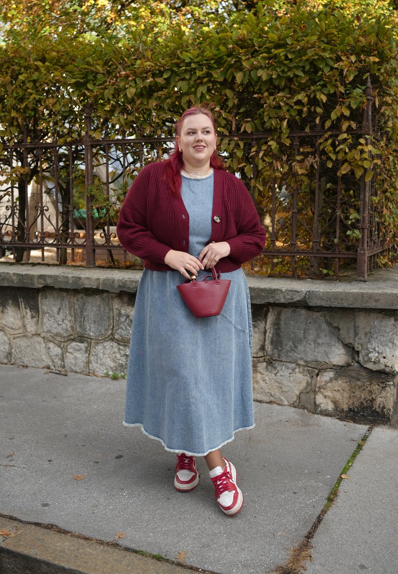 Burgunderfarbener Strickcardigan, hellblaues Jeans-Maxikleid mit fransigem Saum, rot-weiße Turnschuhe und eine kleine rote Handtasche in der Hand.