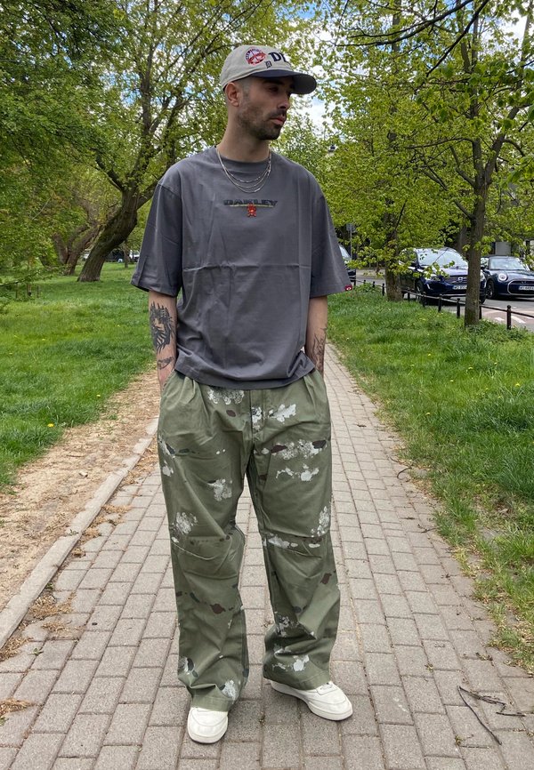Bărbat purtând un tricou gri Oakley, pantaloni de camuflaj, adidași albi și o șapcă albă, stând pe o potecă de cărămidă într-un parc verde.