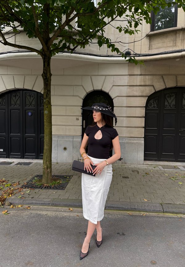 Frau mit schwarzem Hut und Oberteil sowie weißem Rock steht auf der Straße neben einem Baum und schwarzen Türen und hält eine schwarze Clutch mit weißen Punkten.
