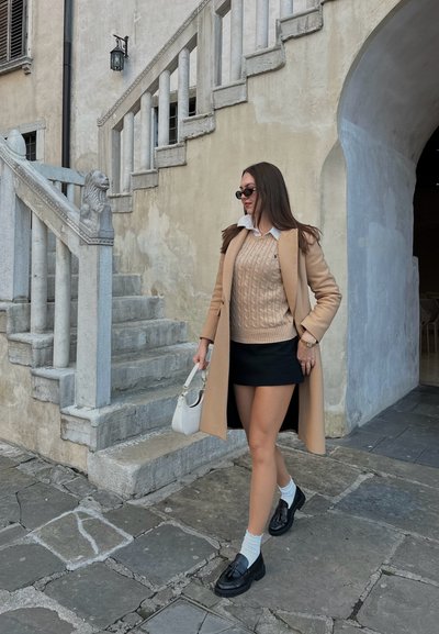 Abrigo beige sobre un suéter de punto de cable, falda corta negra, bolso blanco y zapatos negros con detalle de lazo. De pie junto a una escalera de piedra.