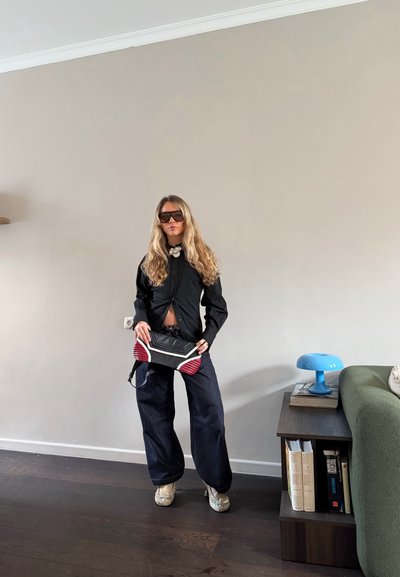 Mujer con cabello largo y rubio, con gafas de sol, camisa negra, pantalones anchos oscuros, sosteniendo un bolso de mano negro y rojo, de pie en el interior junto a una mesa y un sofá.