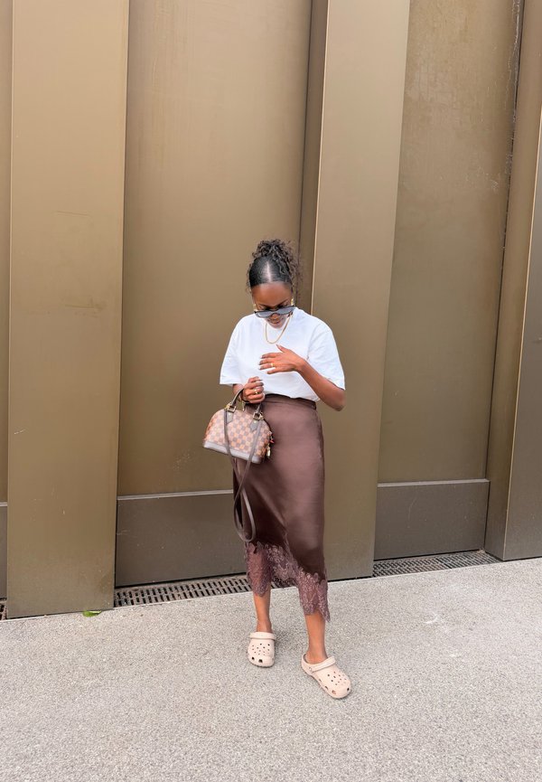 Frau mit lockigem Haar in weissem Hemd und braunem Rock, die eine kariert Handtasche hält, trägt Sonnenbrillen und beige Slip-On-Schuhe.