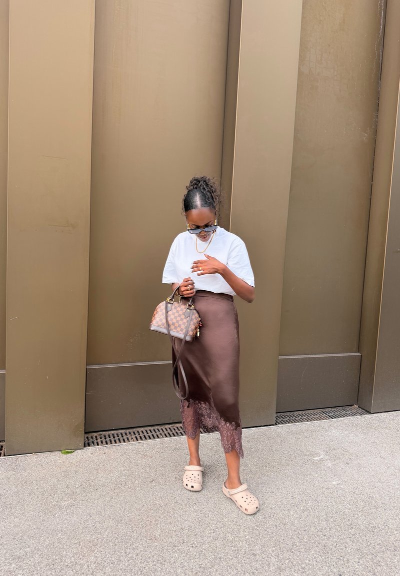 Femme aux cheveux bouclés, portant une chemise blanche et une jupe marron, tenant un sac à main à carreaux, portant des lunettes de soleil et des chaussures beige sans lacets.