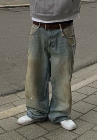 Φαρδιές ανοιχτόμπλε τζιν με ξεθωριασμένα σημεία, ορατές ραφές και φθαρμένες λεπτομέρειες. Φοριούνται με γκρι φούτερ και λευκά αθλητικά παπούτσια.