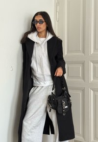 Lange zwarte jas over een grijze hoodie en een joggingbroek. Zwarte handtas met metalen accenten. Model draagt donkere zonnebril. Minimalistische achtergrond.