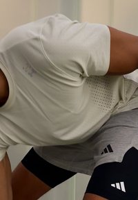 Personne athlétique se penchant en avant, portant une chemise beige perforée, un short gris et des leggings de compression noirs avec des logos blancs.