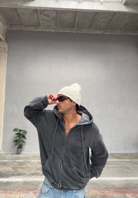 Šedá mikina na zip s kapucí se stahovací šňůrkou a světlejší šedou podšívkou. Nošena s krémovou čepicí a tmavými slunečními brýlemi v industriálním prostředí.