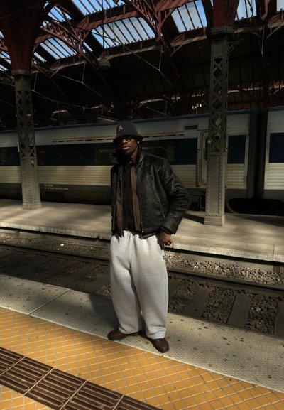 Hombre con chaqueta negra, sombrero tipo pescador, gafas de sol y pantalones deportivos grises de pie en la plataforma de la estación de tren junto a las vías y el tren.