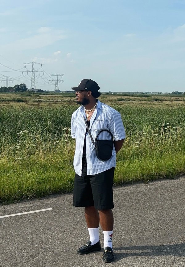 Homem vestido com uma camisa listrada azul e branca, calções pretos, boné preto e sandálias. Ele tem uma pequena bolsa de ombro e meias brancas.