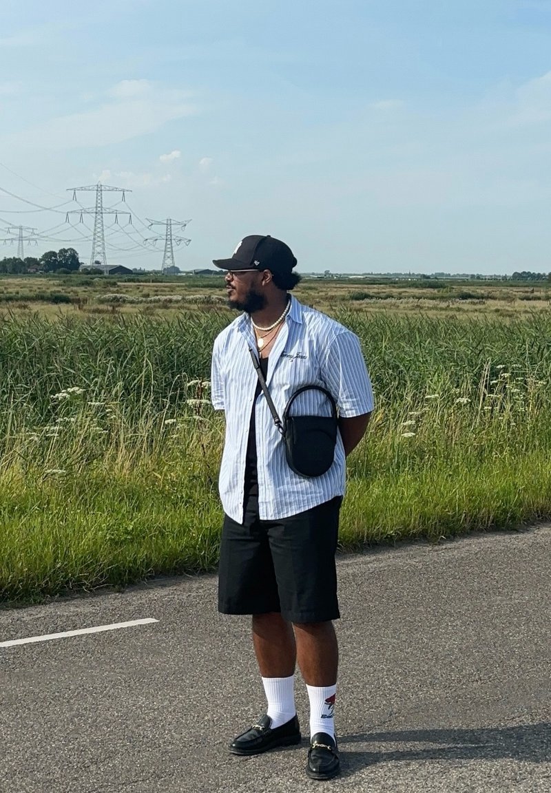 Άνδρας φορώντας ένα μπλε και λευκό ριγέ πουκάμισο, μαύρες βερμούδες, μαύρο καπέλο και σανδάλια. Έχει μια μικρή τσάντα ώμου και λευκές κάλτσες.