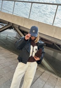 Črna usnjena kratica jakna z velikim ovratnikom, rjava kapuca z belim številom '08', smetana široke hlače in denim kapa. Drži rjavi čevelj.