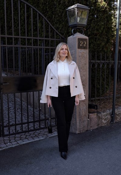 Trinchado beige claro con botones de doble botonadura, usado sobre un suéter blanco de canalé. Jeans negros de tiro alto y zapatos negros puntiagudos.