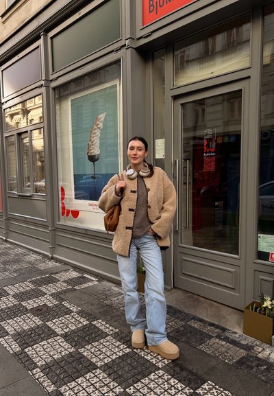 Chaqueta beige de felpa con botones negros, suéter gris, jeans azul claro y zapatos beige sin cordones. Usando auriculares grandes, de pie afuera de una tienda.