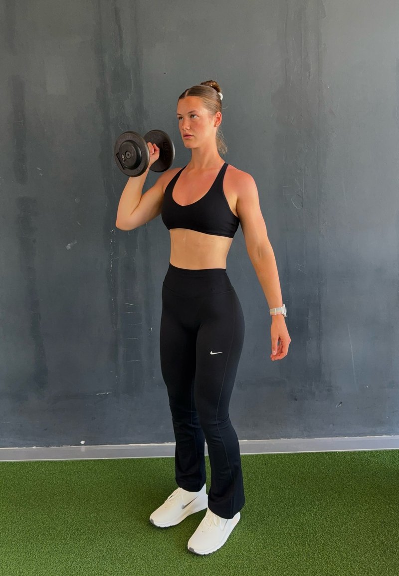 Femme en tenue de sport noire soulevant un haltère près d’un mur gris sur un sol vert, concentrée sur l’entraînement de force avec des baskets blanches et une montre connectée.