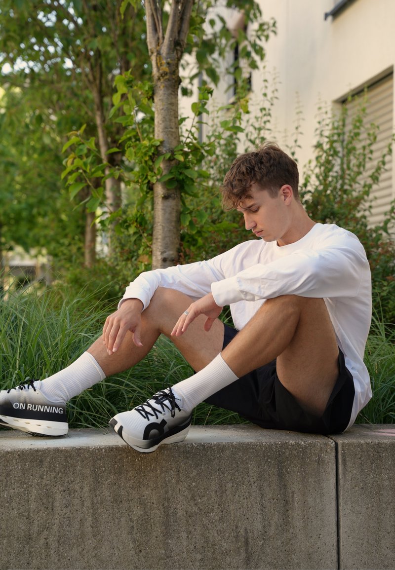 Jeune homme en chemise blanche à manches longues, short noir et chaussures de course blanches assis sur un rebord en béton à l'extérieur près des arbres et de l'herbe.