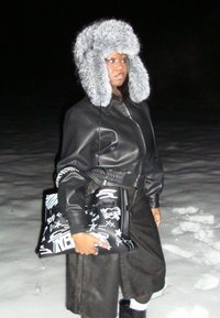 Personne portant une veste en cuir noire et une longue jupe tient un sac fourre-tout noir à imprimé graffiti, coiffée d'un grand chapeau de fourrure gris, debout sur la neige la nuit.