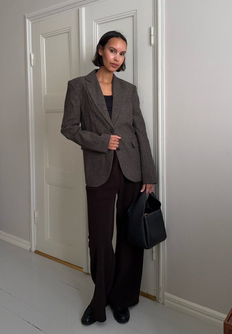 Femme portant un blazer marron oversize et un pantalon à jambes larges, tenant un sac à main noir, debout à l'intérieur près d'une porte blanche fermée.