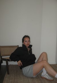 Μαύρο τζάκετ με φερμουάρ, γκρι σόρτς, λευκές κάλτσες και γκρι αθλητικά παπούτσια με καφέ λεπτομέρειες. Υφασμάτινη καρέκλα στο φόντο. Ουδέτερο τοίχος.