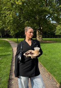 Svart oversized blazer, lyseblå vide jeans, holder et brett med solbriller og iskald kaffe i en parkinnstilling med gress og trær.