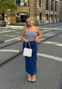 Raidallinen olkaimeton toppi, sininen maksihame ja beige puukengät. Valkoinen teksturoidu käsilaukku, kaupunkitausta kivirakennuksen ja kadun kanssa.