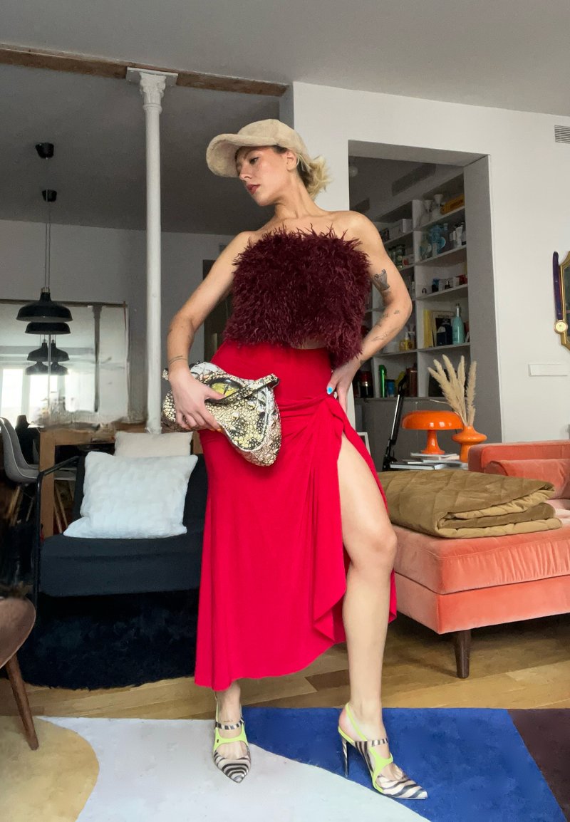 Femme en robe rouge avec fente sur la cuisse, haut bordeaux à plumes, casquette beige, tenant une pochette en peau de serpent, debout dans un salon moderne.