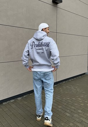 Sudadera con capucha gris con la frase "Prohibited Garments" en cursiva azul marino en la parte trasera, combinada con vaqueros azul claro y zapatillas beige con detalles en negro.