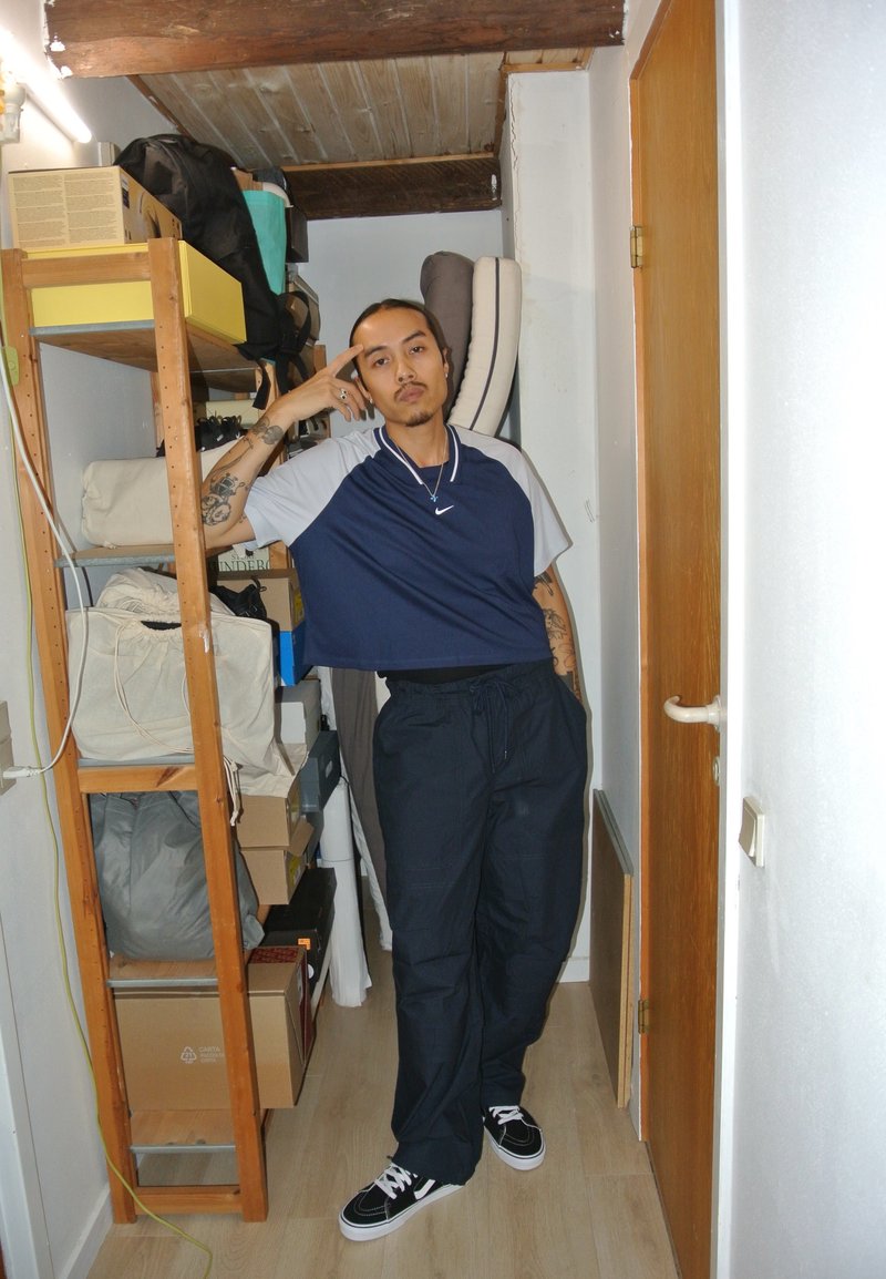 Μπλε και γκρι κοντό αθλητικό μπλουζάκι, μαύρο φαρδύ παντελόνι και μαύρα αθλητικά παπούτσια με λευκές ρίγες. Το φόντο περιλαμβάνει ράφια αποθήκευσης και κουτιά.