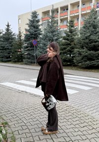 Bruine oversized jas, bijpassende bruine broek en een bruine en witte handtas met patroon. Staand op een bakstenen pad met altijdgroene bomen op de achtergrond.