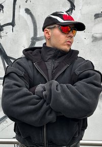 Giacca nera oversize con zip, foderata in pile, collo alto e ampi polsini. Cappello nero, occhiali da sole specchiati e dettagli in argento sugli orecchini.