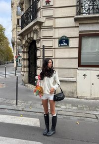 Mustad kummist vihmataldad, valge kootud kampsun ja valged lühikesed püksid. Hoides lillekimbu, õlal must käekott.