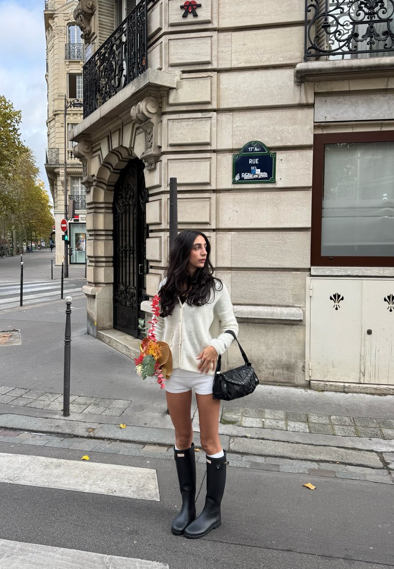 Botas de chuva de borracha preta, suéter branco de malha e calções brancos. Segurando um arranjo floral, com uma mala preta ao ombro.