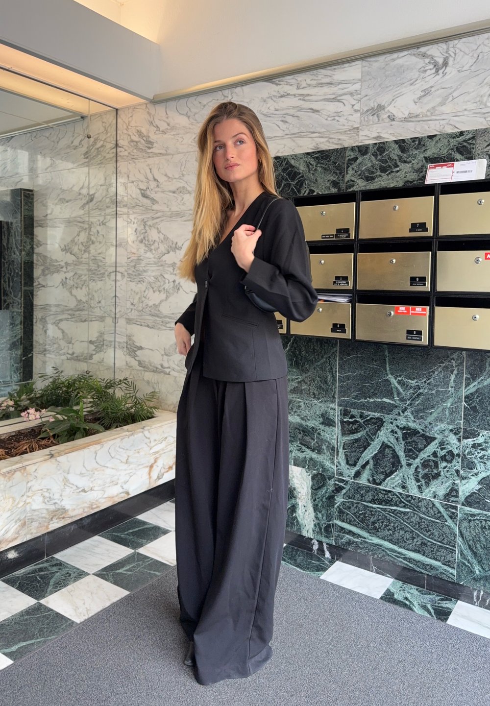 Veste noire ajustée avec manches longues et fermeture à boutons sur le devant, associée à un pantalon noir à jambes larges. Tissu lisse, design minimaliste.