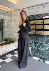 Veste noire ajustée avec manches longues et fermeture à boutons sur le devant, associée à un pantalon noir à jambes larges. Tissu lisse, design minimaliste.