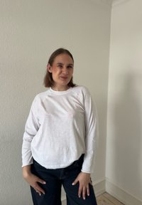 Femme aux cheveux bruns mi-longs portant un haut blanc à manches longues et un pantalon foncé, debout devant un mur uni de couleur claire.
