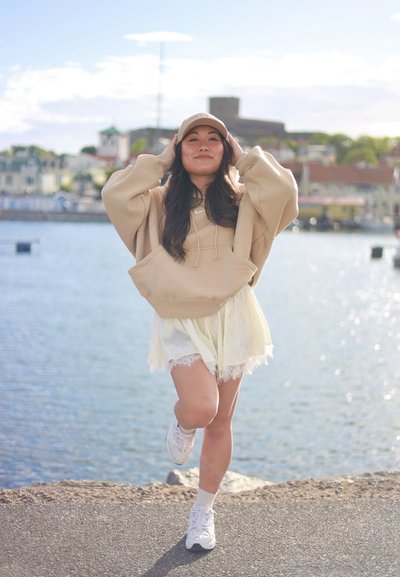 Mujer con sudadera beige con capucha y gorra, equilibrándose sobre una pierna junto al agua con edificios y un castillo al fondo en un día soleado.