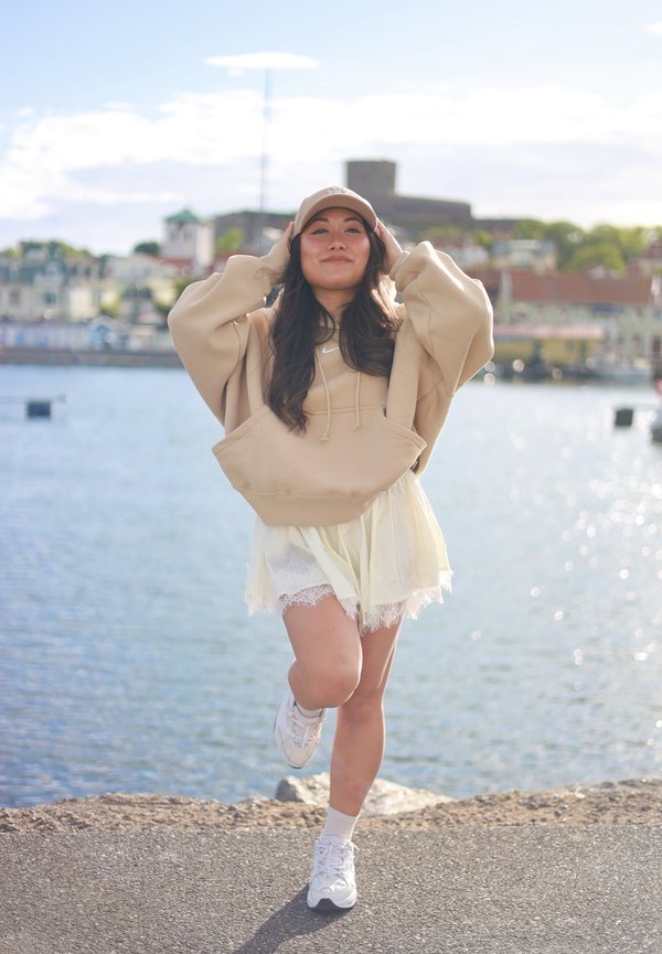 Femme portant un sweat à capuche beige et une casquette, en équilibre sur une jambe au bord de l'eau avec des bâtiments et un château en arrière-plan par une journée ensoleillée.